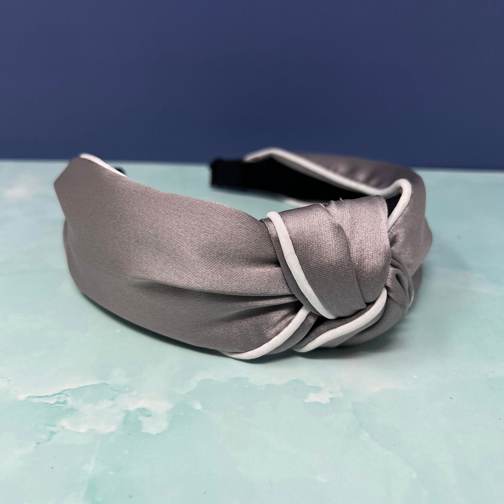 Gray satin knotted headband with white piping, displayed on a soft blue background.