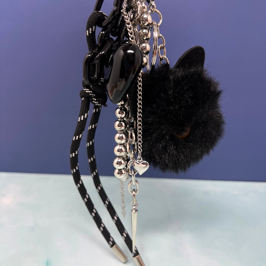 Black cat plush charm keychain with silver chain cluster, black heart, and spike pendant clipped onto hand.