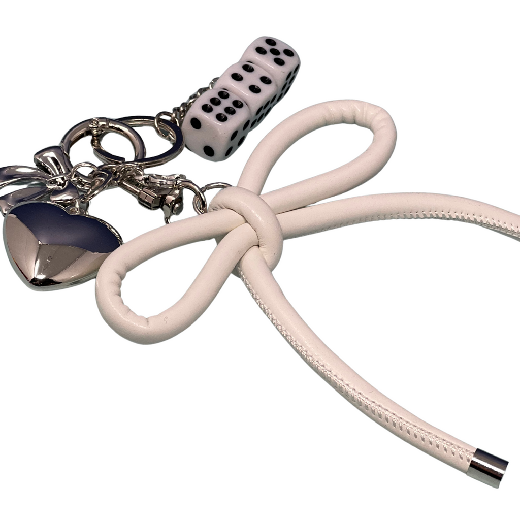 White faux leather bow keychain with silver heart charm, bow accent, and stacked dice charms on a blue surface