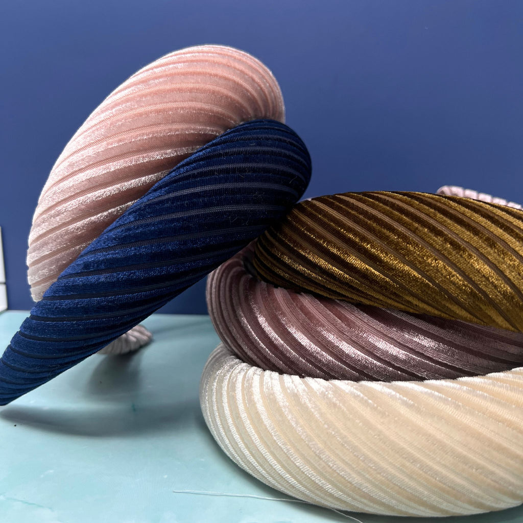 Assorted pleated velvet padded headbands in navy, mauve, light pink, brown, cream, and burgundy stacked in a colorful display.