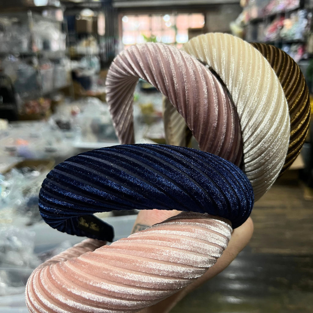 Assorted pleated velvet padded headbands in navy, mauve, light pink, brown, cream, and burgundy stacked in a colorful display.