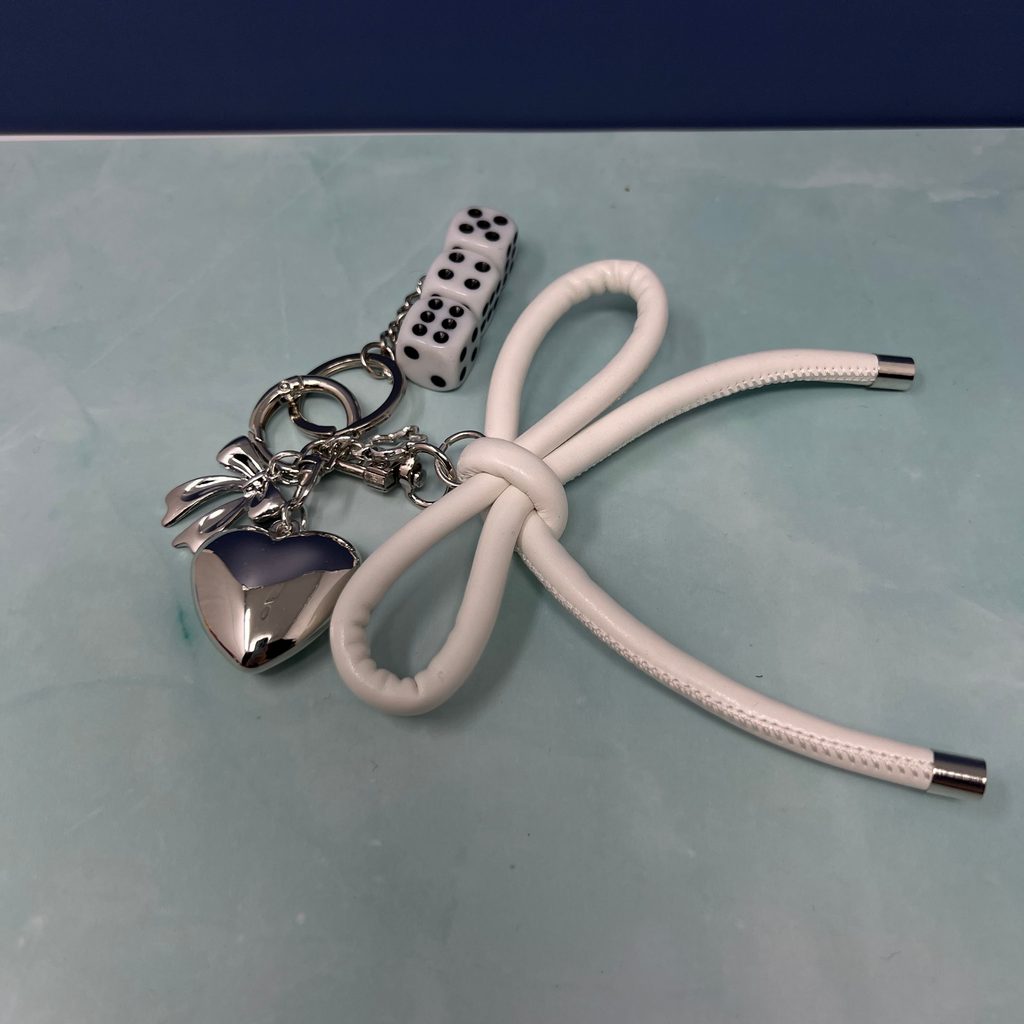White faux leather bow keychain with silver heart charm, bow accent, and stacked dice charms on a blue surface