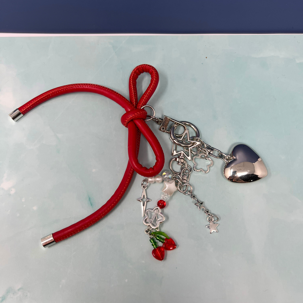 Red faux leather bow keychain with silver heart charm, cherry beads, stars, and flower dangles on light blue background