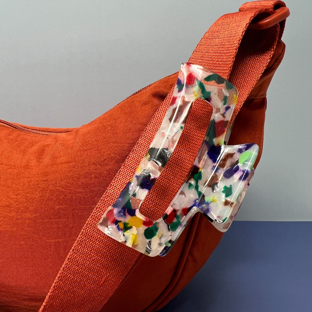 Clear acrylic lightning bolt-shaped hair claw with multicolored confetti design, clipped onto an orange fabric bag strap, styled on a color-blocked background.

