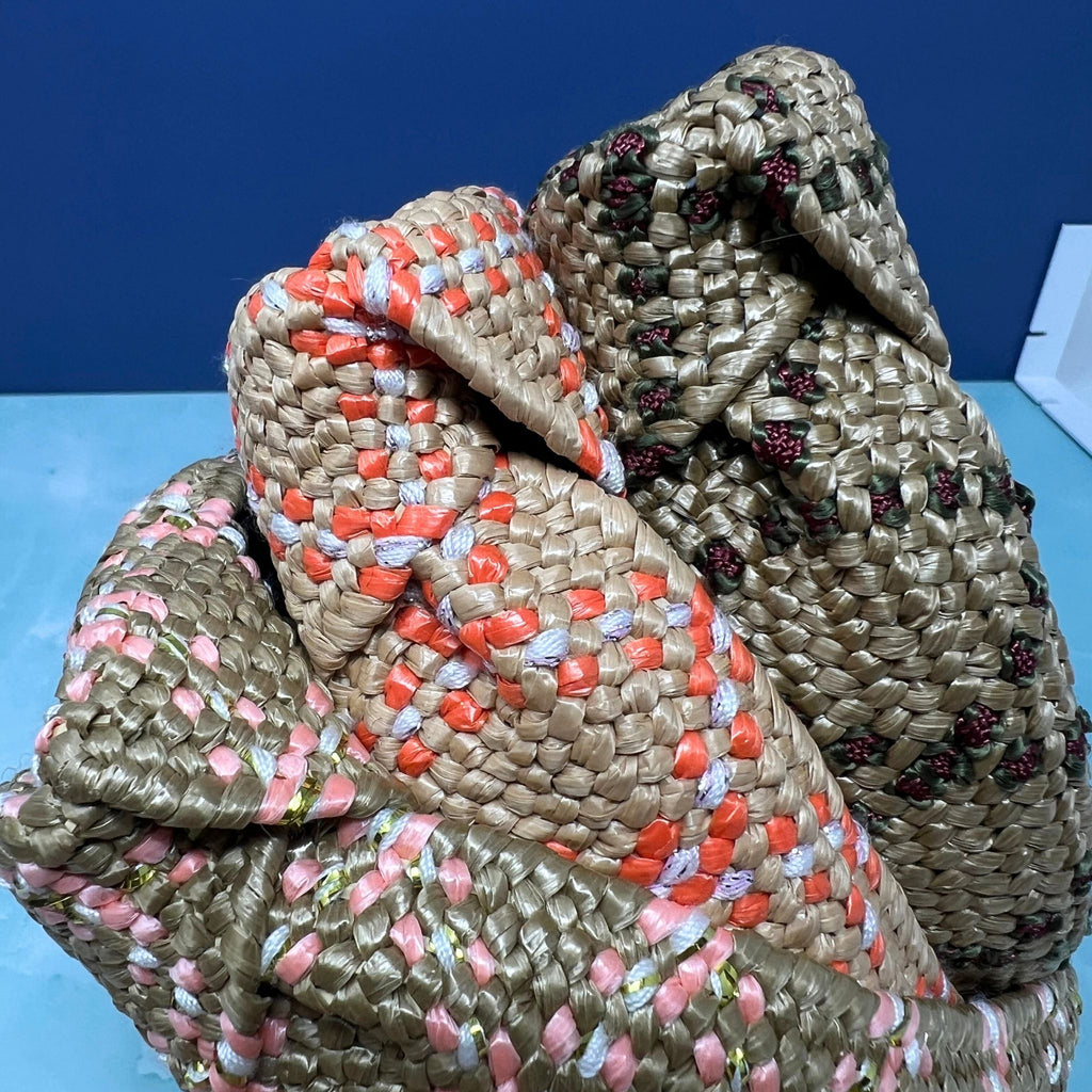 Three braided raffia-style knotted headbands in natural straw color with coral, pink, and floral embroidery accents, stacked against a blue background.