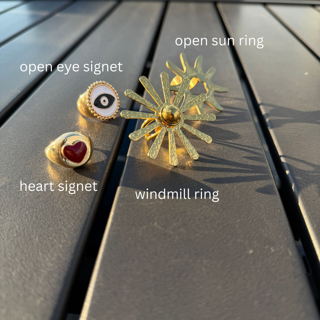Four gold rings labeled 'open eye signet', 'heart signet', 'open sun ring', and 'windmill ring' on a wooden surface.