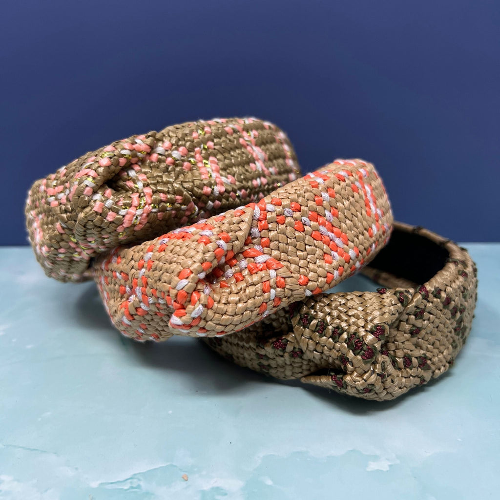 Three braided raffia-style knotted headbands in natural straw color with coral, pink, and floral embroidery accents, stacked against a blue background.