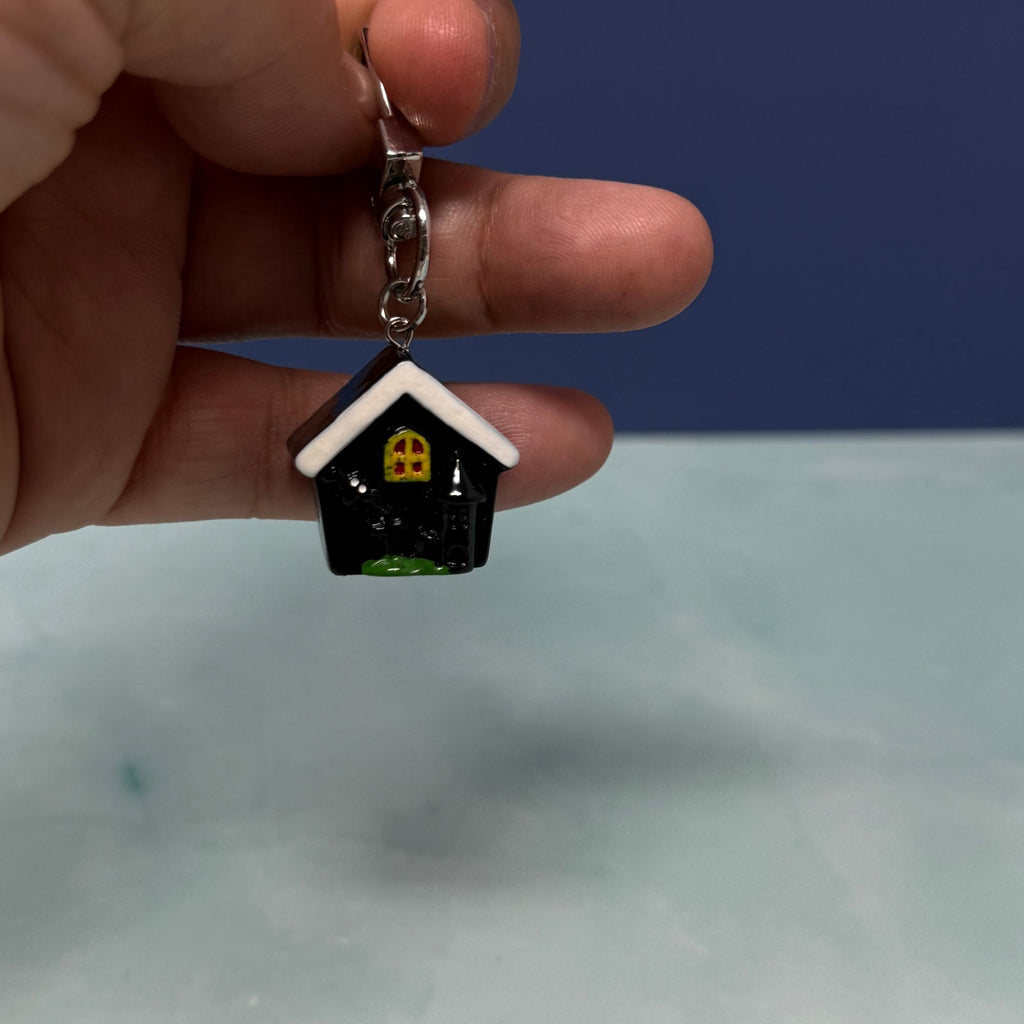 Tiny black and white house mini keychain with yellow window, glossy finish, and silver clasp held in hand.