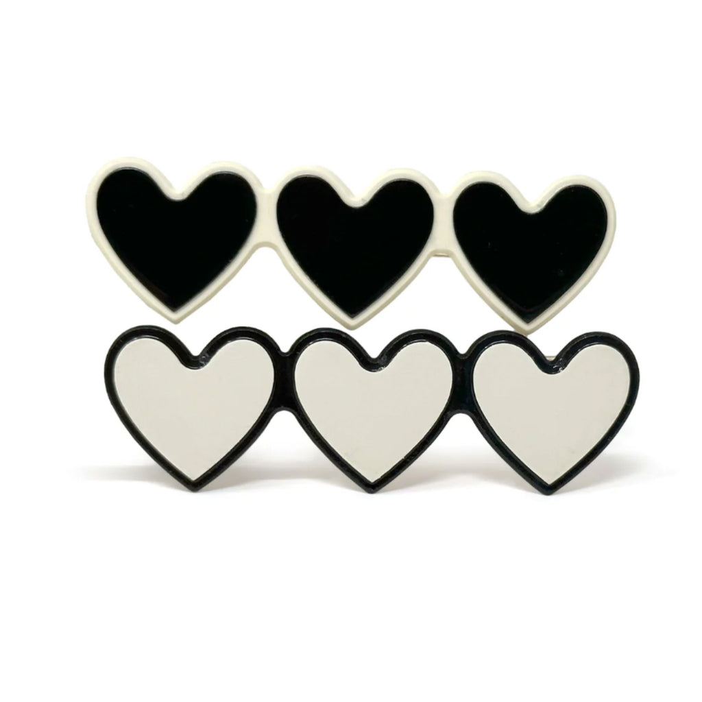 Set of two triple-heart barrettes in black with white outline and white with black outline, each measuring approximately 2.5"W x 0.75"H with secure alligator clip backing.