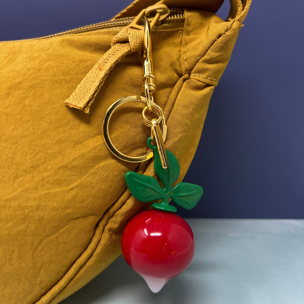 Radish keychain with glossy red root, white tip, green enamel leaves, and gold hardware displayed against a light background