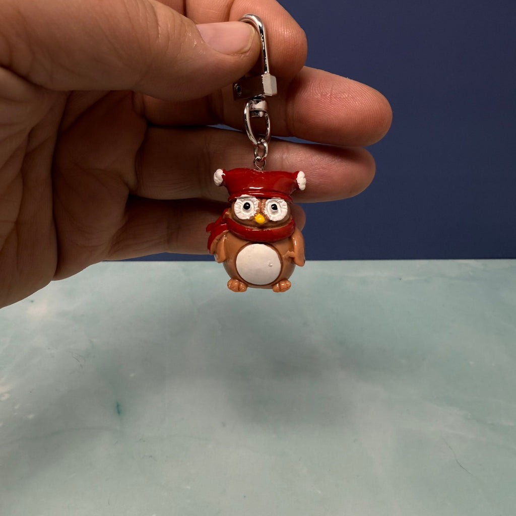 Mini owl keychain wearing a red winter hat and scarf, held in hand against a blue background.