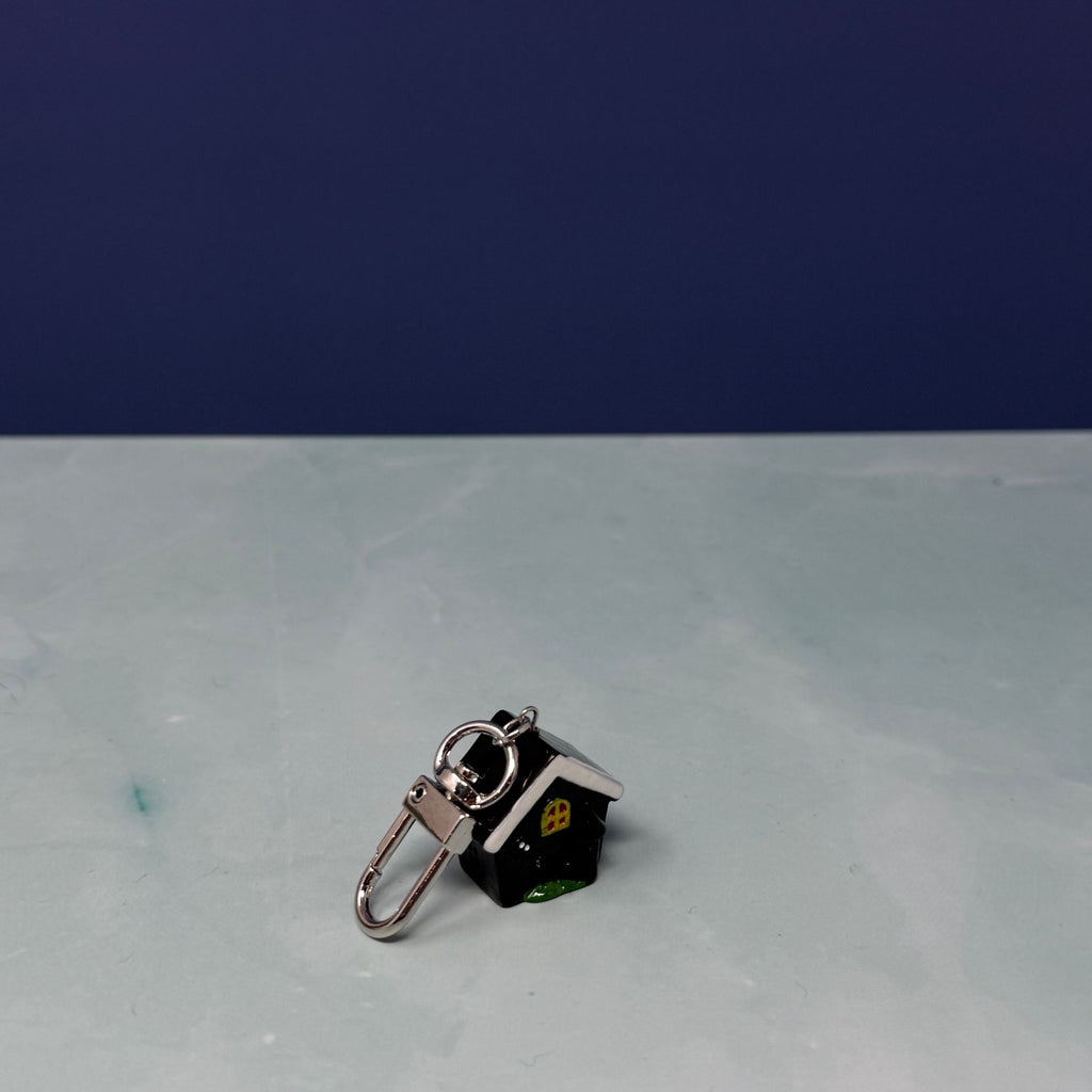 Tiny black and white house mini keychain with yellow window, glossy finish, and silver clasp held in hand.