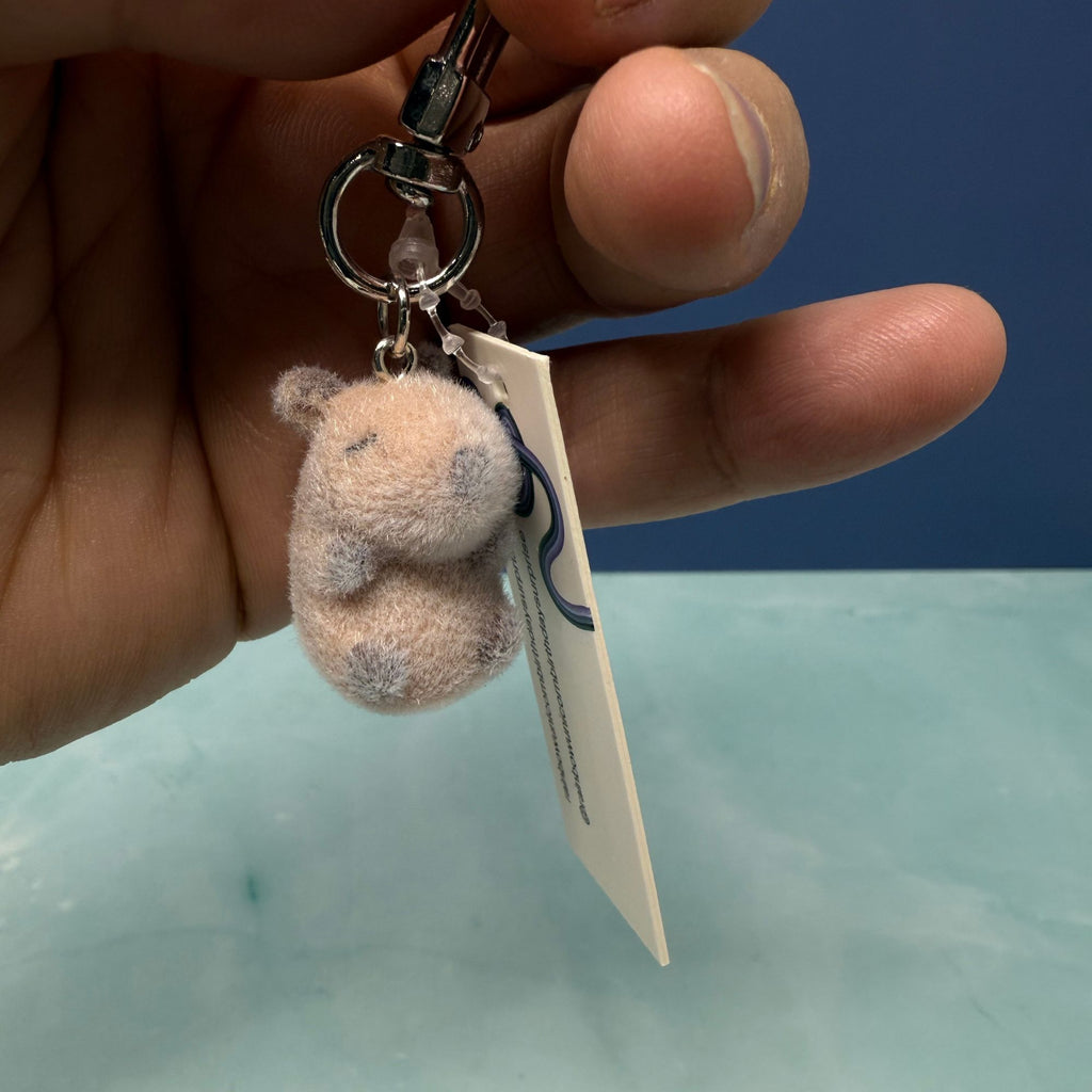 Flocked capybara mini plush keychain with sleepy face held by hand on blue background