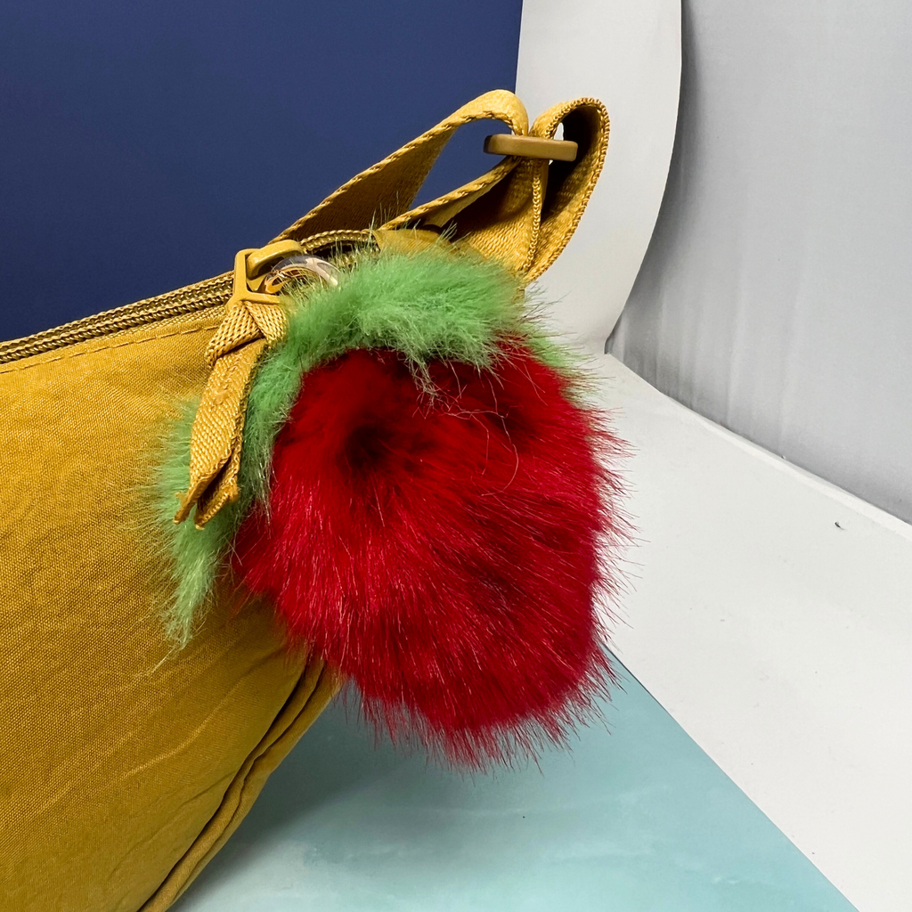 Plush red strawberry pom pom keychain with green fuzzy leaf top, clipped onto a mustard yellow handbag.