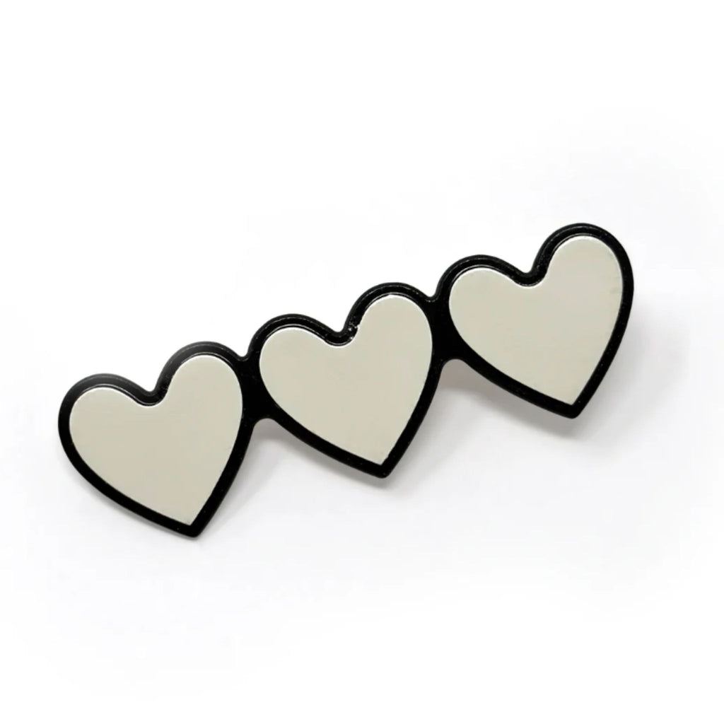 Set of two triple-heart barrettes in black with white outline and white with black outline, each measuring approximately 2.5"W x 0.75"H with secure alligator clip backing.