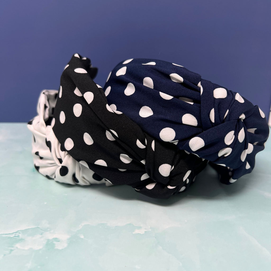 Stack of three polka dot knotted headbands in black, navy, and white with white dots, styled for retro or classic looks.