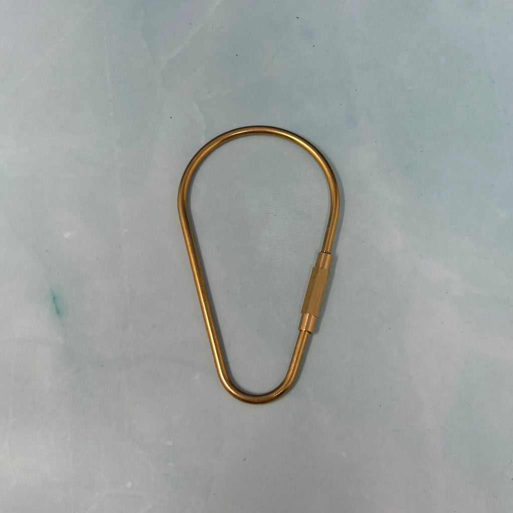 Flat lay of assorted geometric brass key rings including teardrop, oval, rectangle, half-moon, and paperclip shapes on a light blue surface.