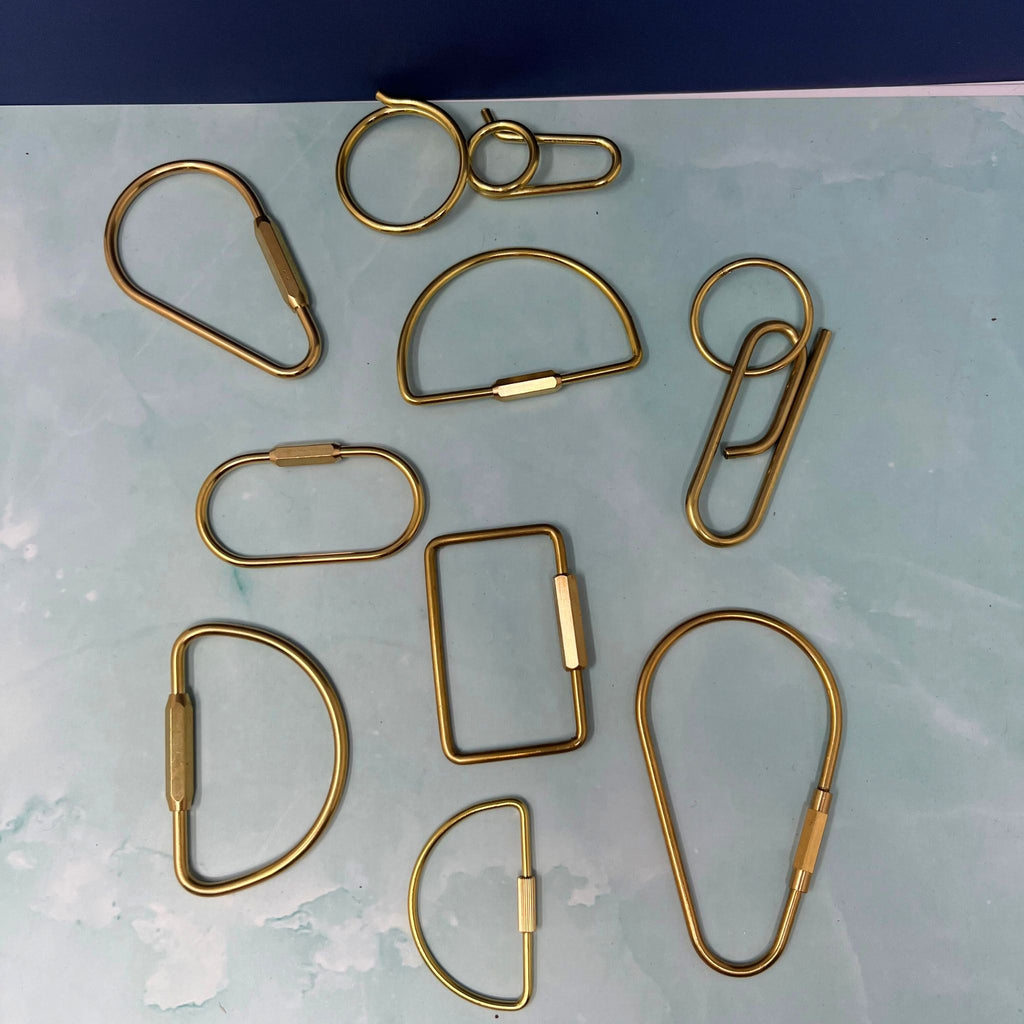 Flat lay of assorted geometric brass key rings including teardrop, oval, rectangle, half-moon, and paperclip shapes on a light blue surface.