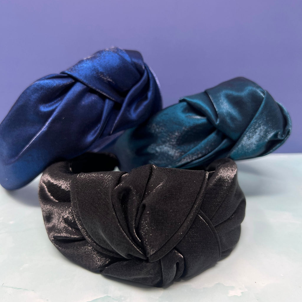 Three shiny satin knot headbands in black, navy, and teal stacked on a flat surface with a soft knotted top design.