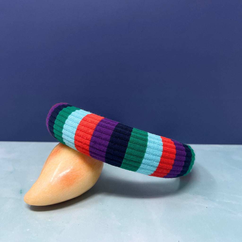 Stack of thick rainbow-striped knit headbands in assorted bold color palettes with one arched to show interior faux suede lining.