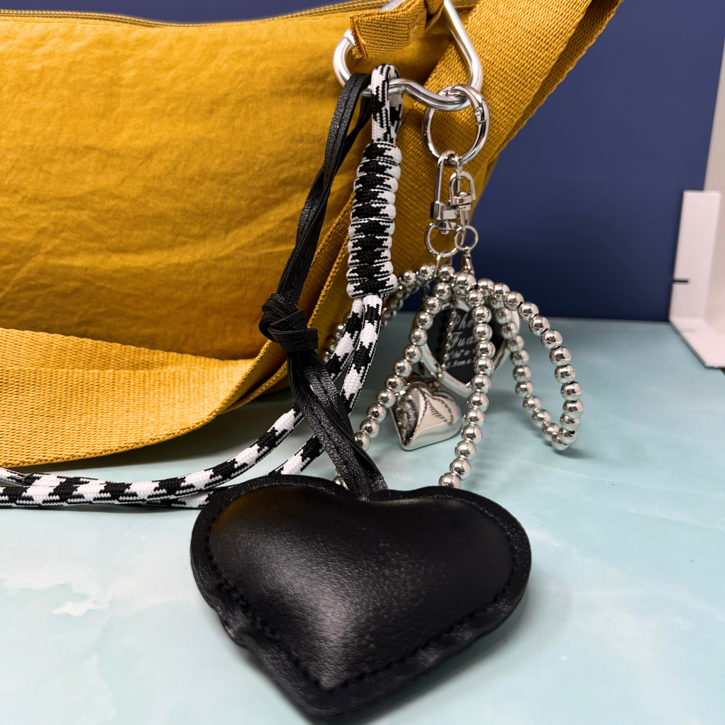 Black faux leather heart keychain with silver charms, pearl bow, and black-and-white rope detail hanging from yellow handbag.