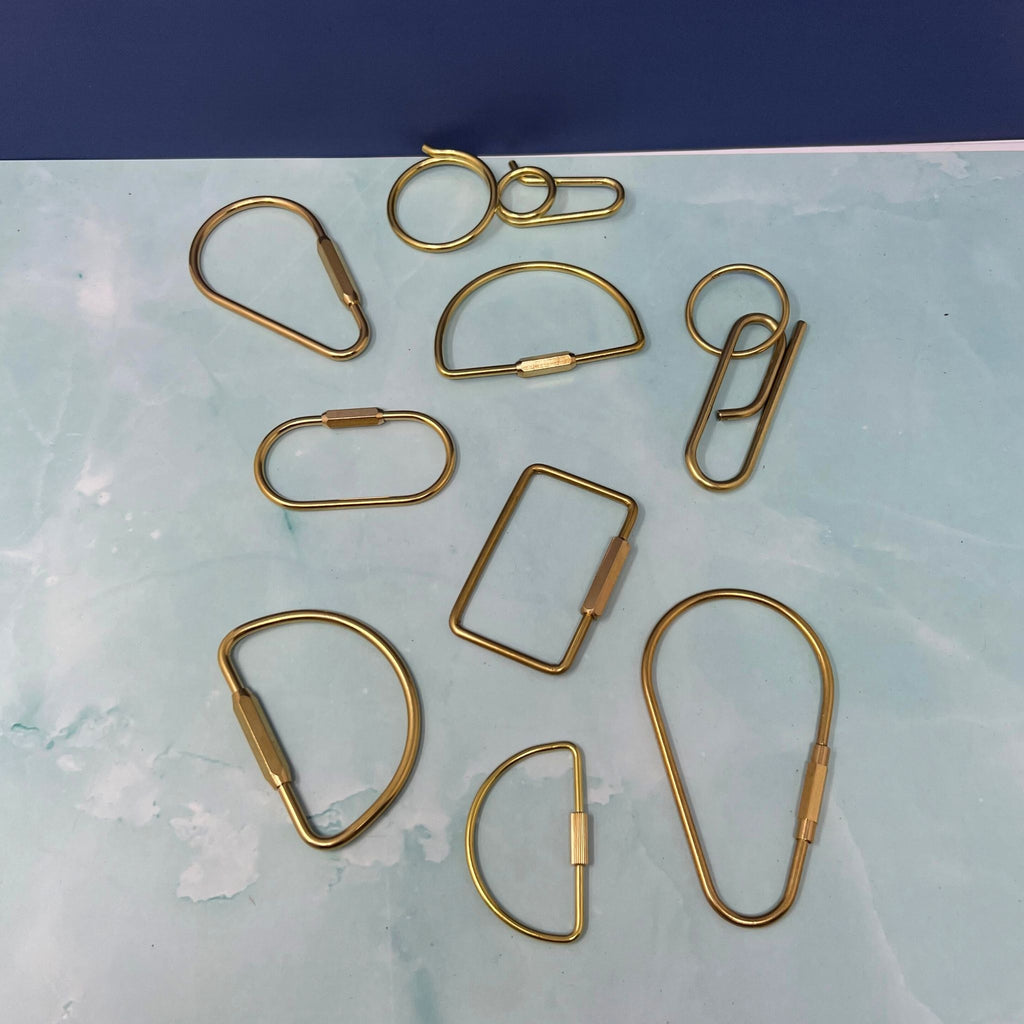Flat lay of assorted geometric brass key rings including teardrop, oval, rectangle, half-moon, and paperclip shapes on a light blue surface.