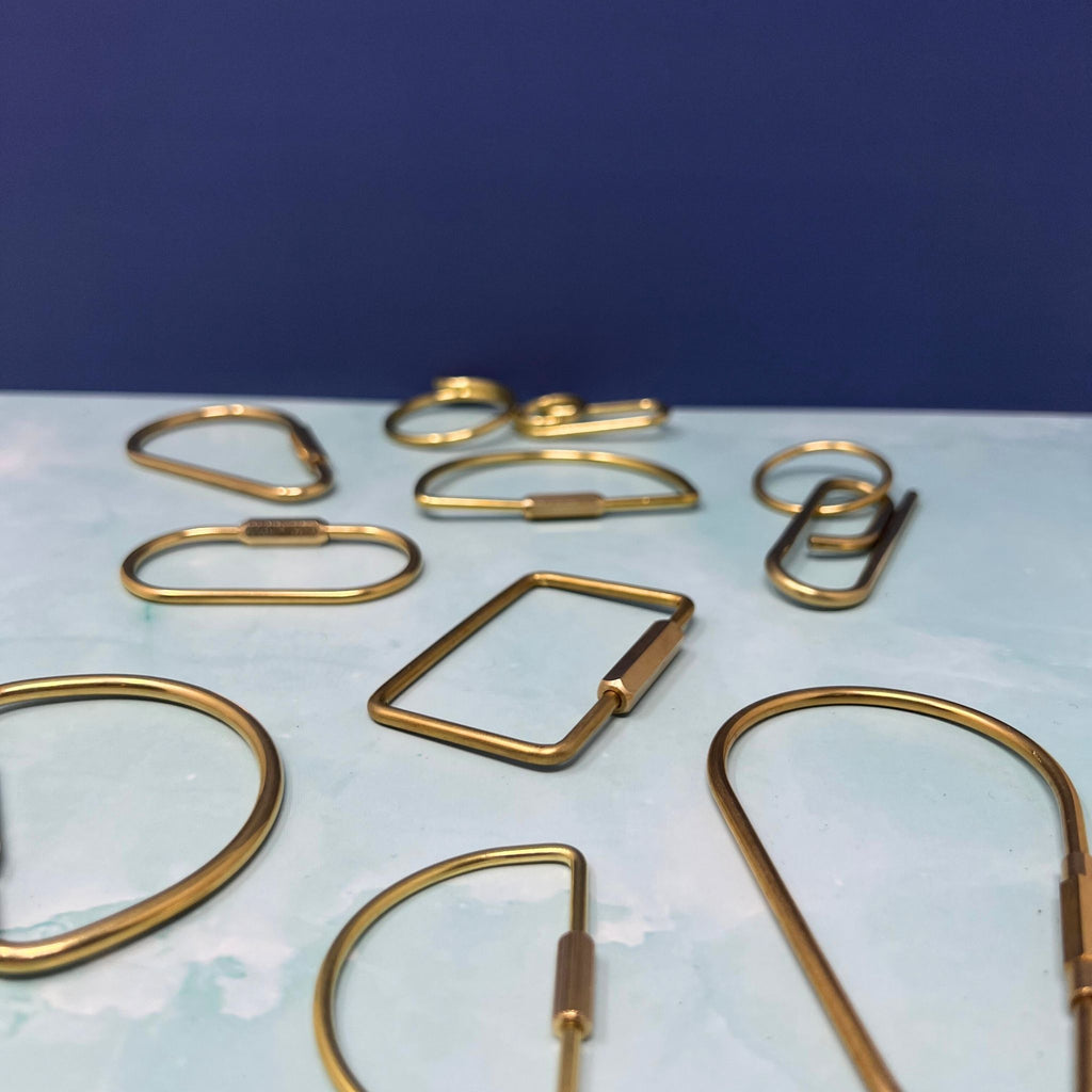 Flat lay of assorted geometric brass key rings including teardrop, oval, rectangle, half-moon, and paperclip shapes on a light blue surface.