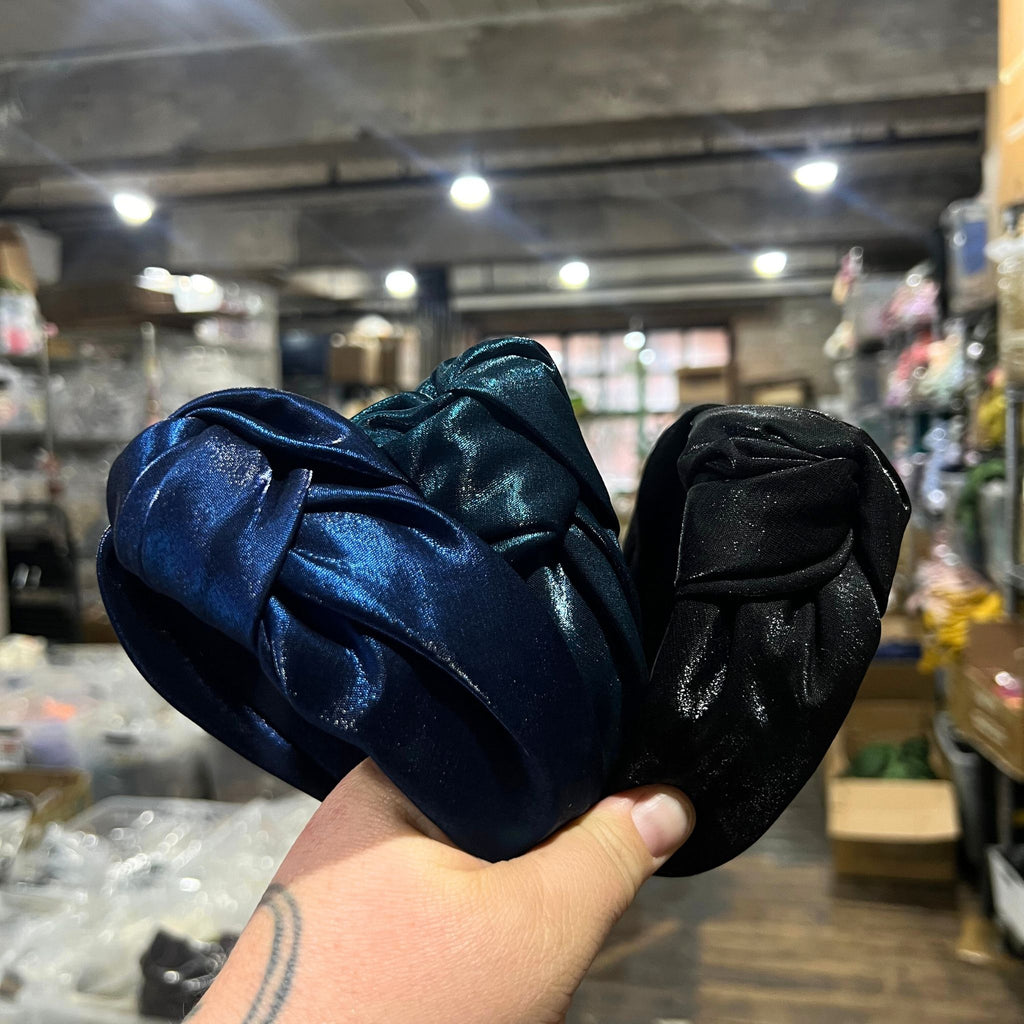 Three shiny satin knot headbands in black, navy, and teal stacked on a flat surface with a soft knotted top design.