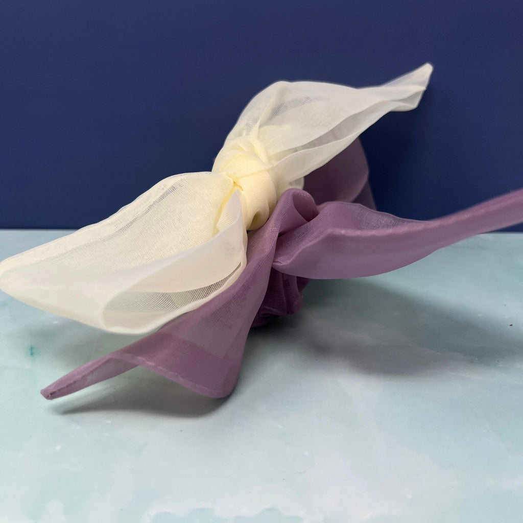 Sheer organza bow scrunchie in cream and lavender with oversized flowing tails, styled on a pastel background.


