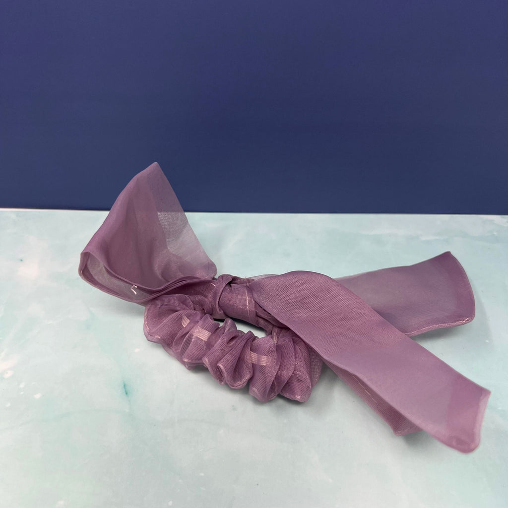Sheer organza bow scrunchie in cream and lavender with oversized flowing tails, styled on a pastel background.

