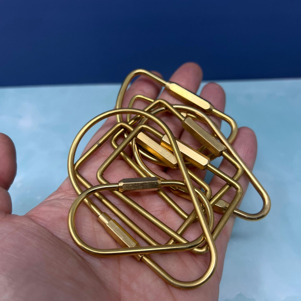 Flat lay of assorted geometric brass key rings including teardrop, oval, rectangle, half-moon, and paperclip shapes on a light blue surface.