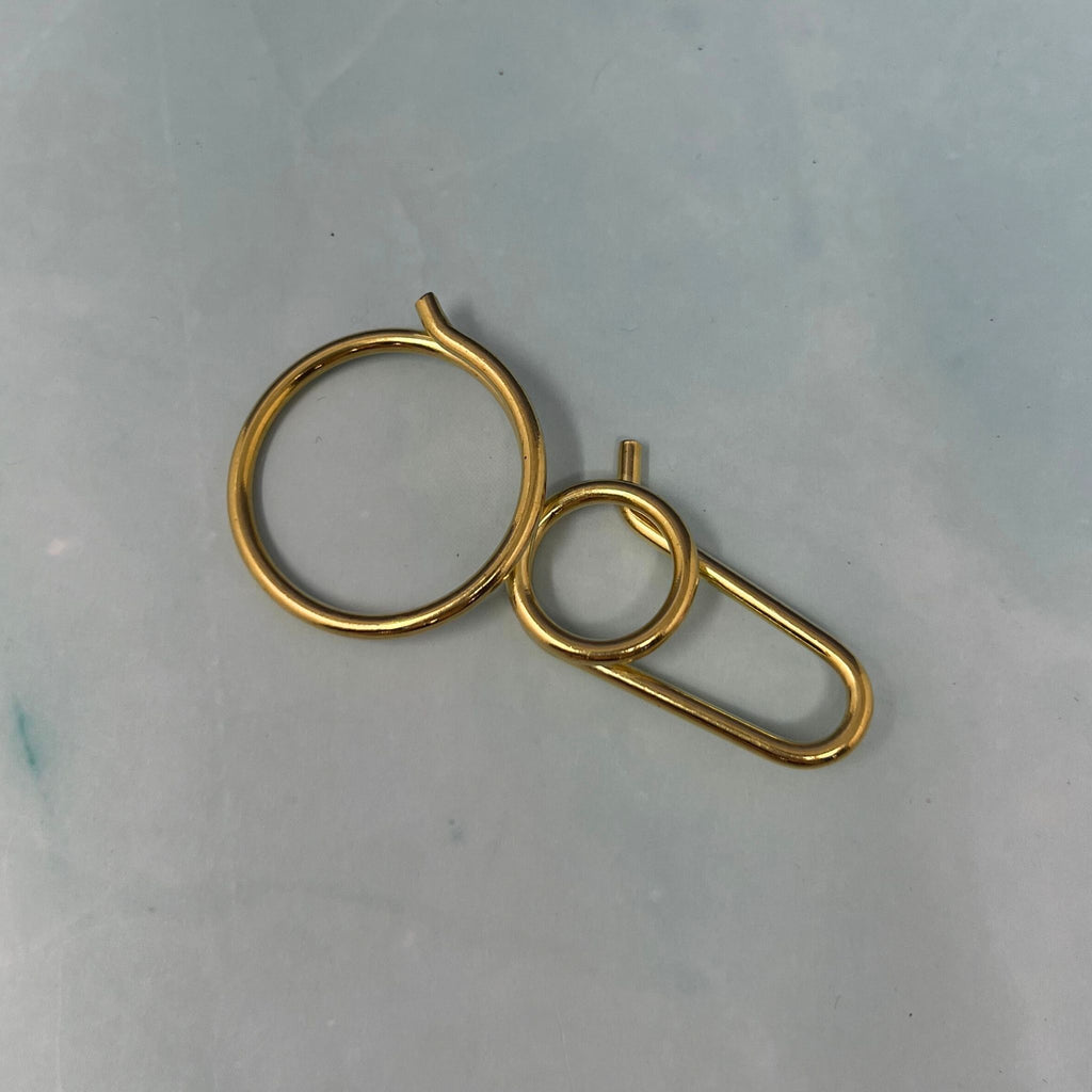 Flat lay of assorted geometric brass key rings including teardrop, oval, rectangle, half-moon, and paperclip shapes on a light blue surface.