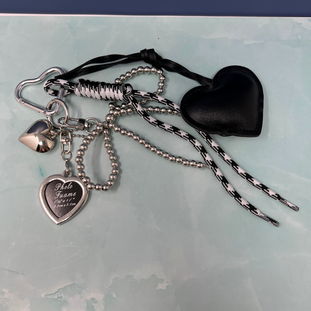 Black faux leather heart keychain with silver charms, pearl bow, and black-and-white rope detail hanging from yellow handbag.