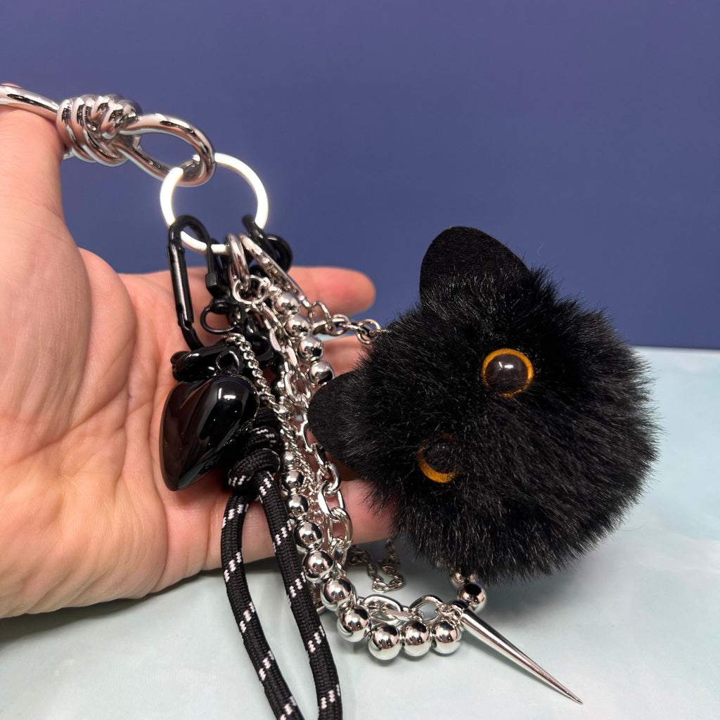Black cat plush charm keychain with silver chain cluster, black heart, and spike pendant clipped onto hand.