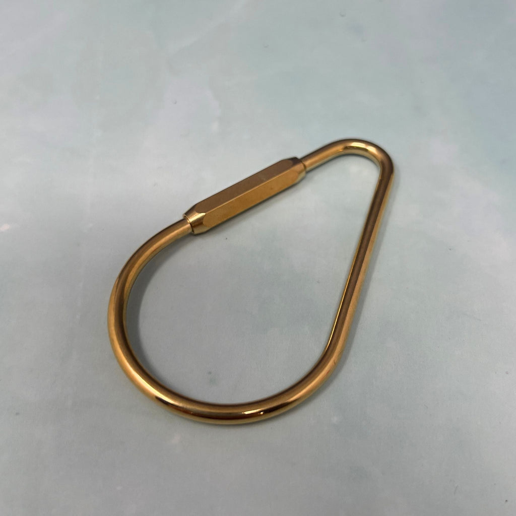 Flat lay of assorted geometric brass key rings including teardrop, oval, rectangle, half-moon, and paperclip shapes on a light blue surface.