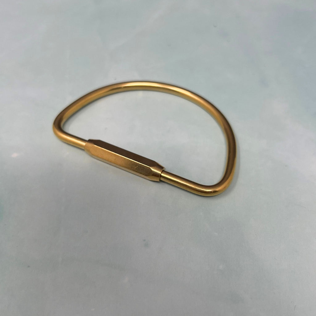 Flat lay of assorted geometric brass key rings including teardrop, oval, rectangle, half-moon, and paperclip shapes on a light blue surface.