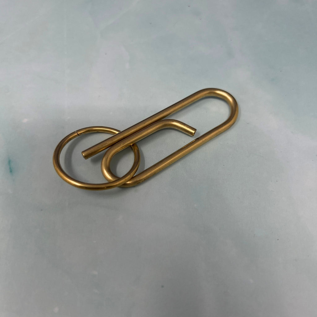 Flat lay of assorted geometric brass key rings including teardrop, oval, rectangle, half-moon, and paperclip shapes on a light blue surface.