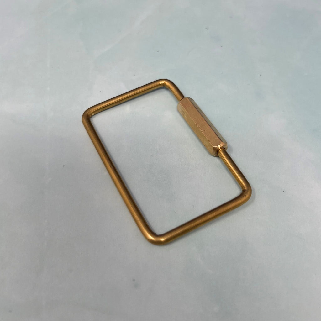 Flat lay of assorted geometric brass key rings including teardrop, oval, rectangle, half-moon, and paperclip shapes on a light blue surface.