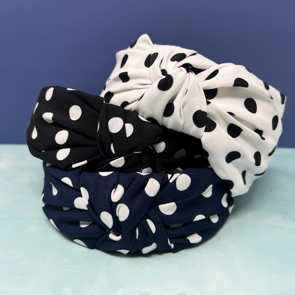 Stack of three polka dot knotted headbands in black, navy, and white with white dots, styled for retro or classic looks.