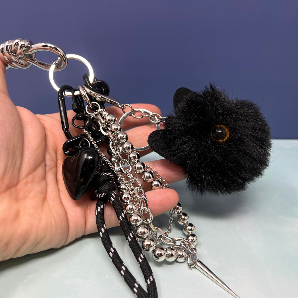 Black cat plush charm keychain with silver chain cluster, black heart, and spike pendant clipped onto hand.