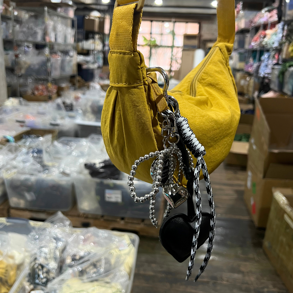 Black faux leather heart keychain with silver charms, pearl bow, and black-and-white rope detail hanging from yellow handbag.