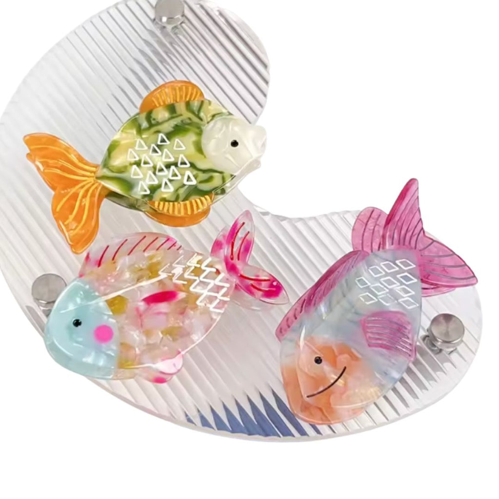Three colorful fish-shaped hair claw clips in iridescent pink, green, and multicolor designs displayed on a clear wavy tray.