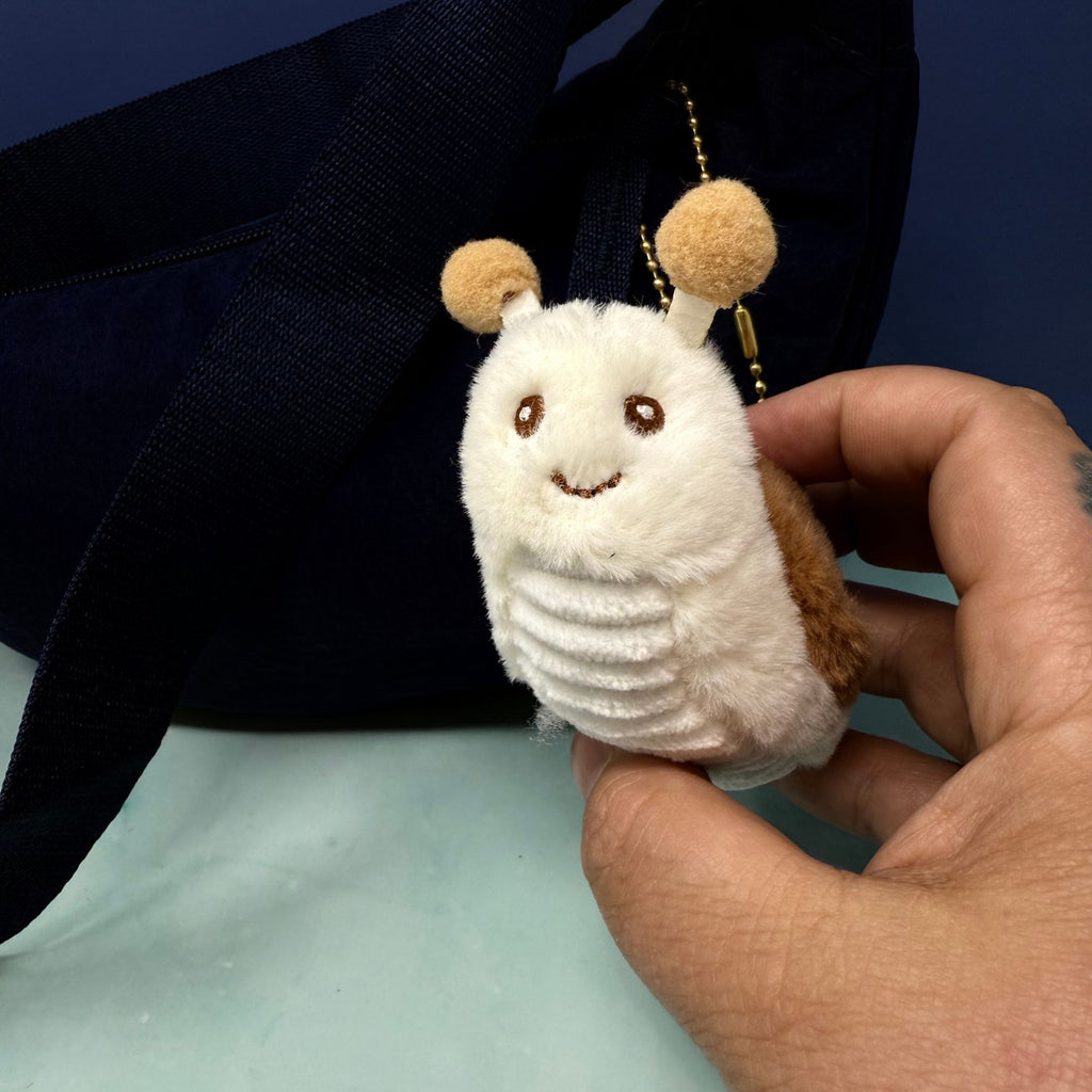 Soft plush snail keychain with tan shell, white body, and smiling face attached to a gold ball chain, shown hanging from a navy bag.
