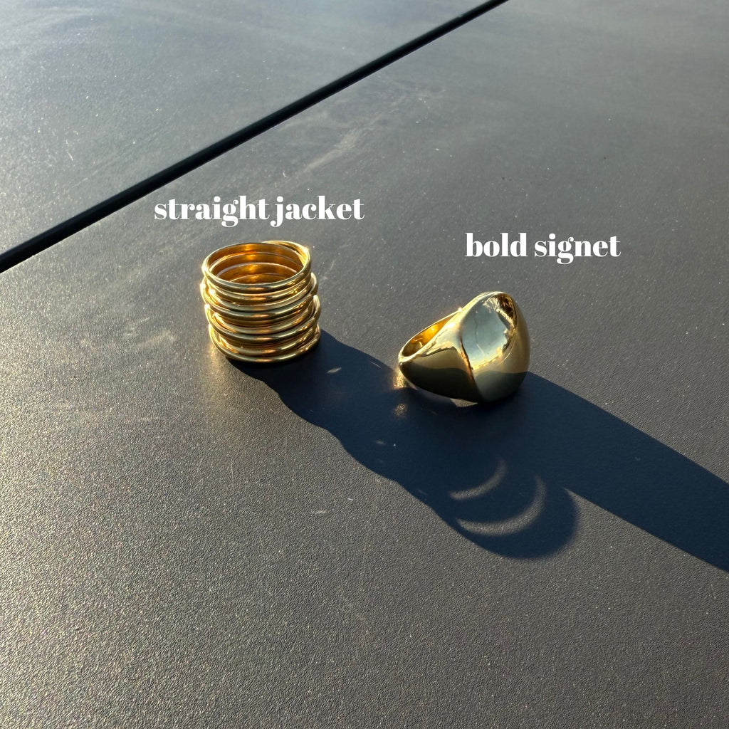 Two gold rings labeled 'straight jacket' and 'bold signet' on a dark surface.