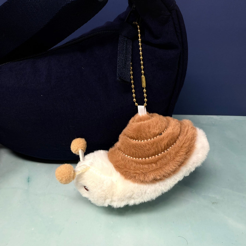 Soft plush snail keychain with tan shell, white body, and smiling face attached to a gold ball chain, shown hanging from a navy bag.