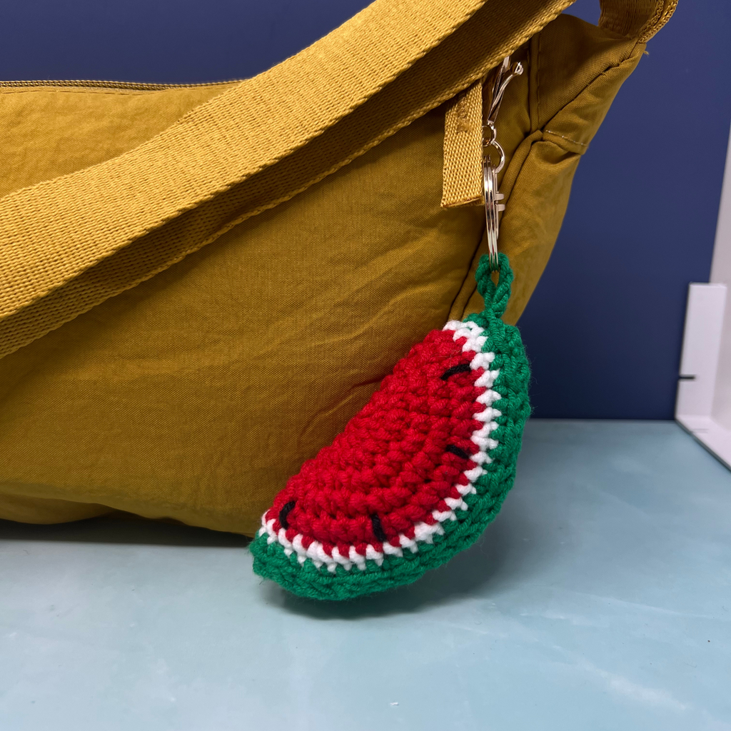 Crochet watermelon slice keychain with gold clasp hanging on a mustard yellow bag.