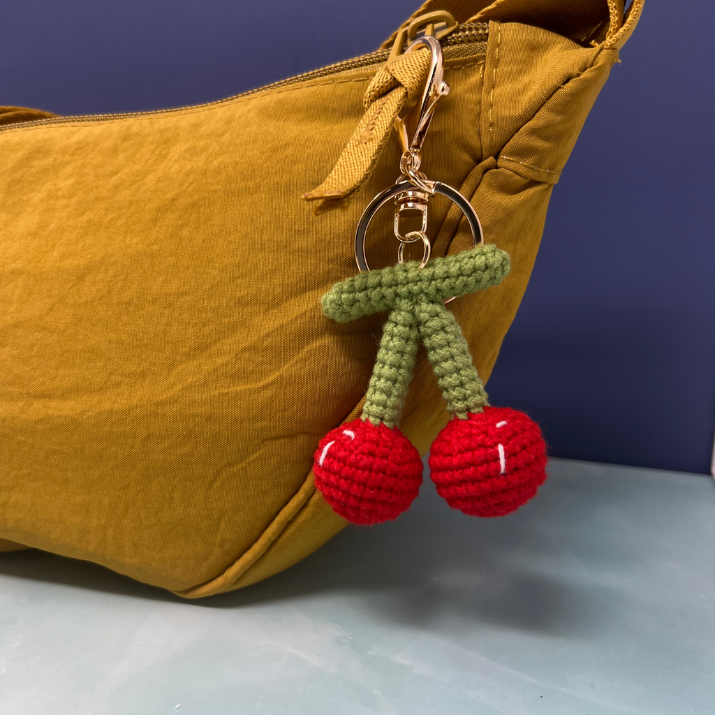 Crochet cherry keychain with red cherries and green stems attached to a gold clip on a mustard bag.